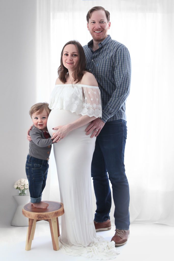 Son stands on stool next to dad and pregnant mom in white room during child and maternity photoshoot