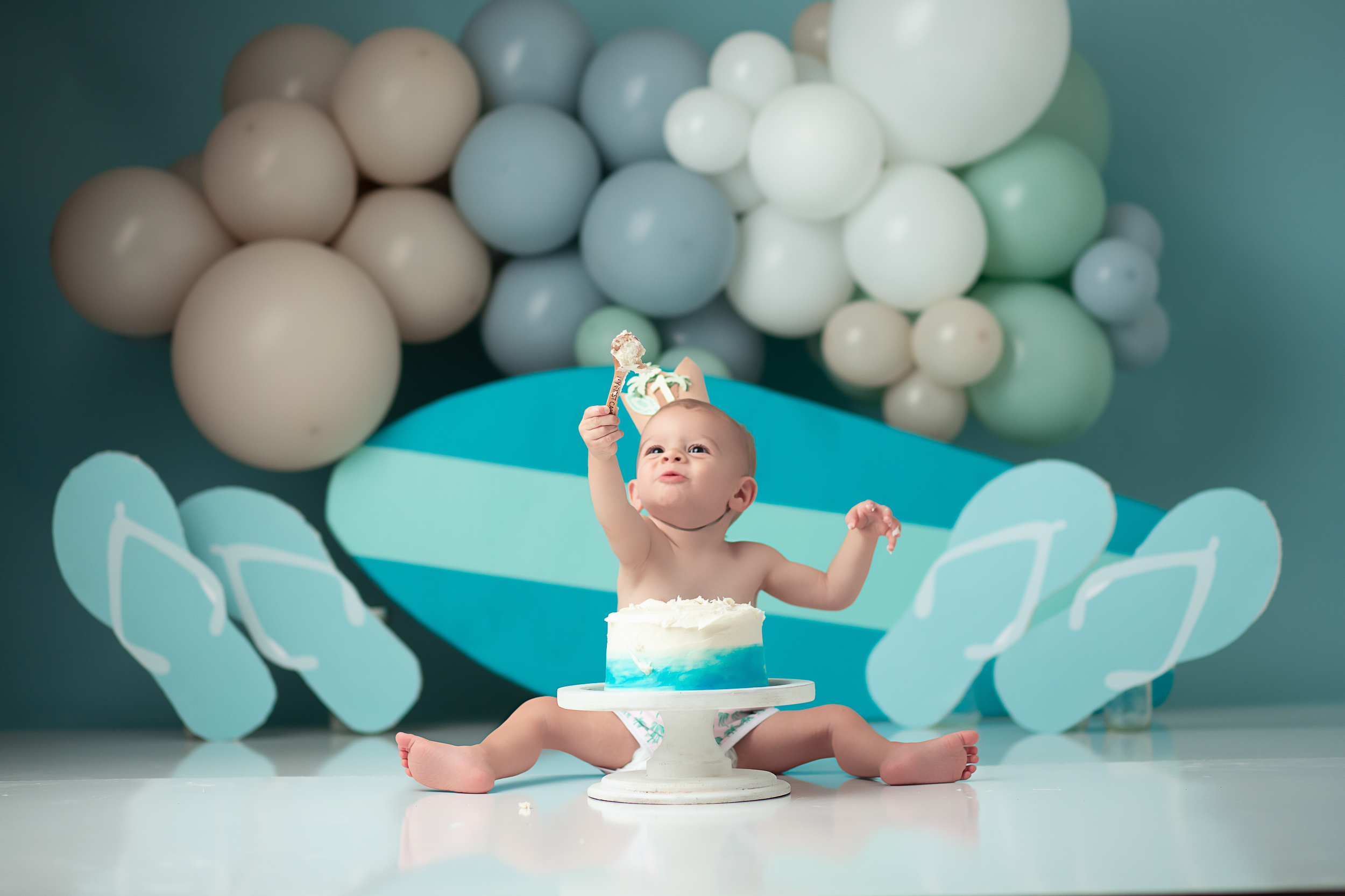 Baby Boy with a cake for a year session with a backdrop of a surfboard, flip flops and balloon banner