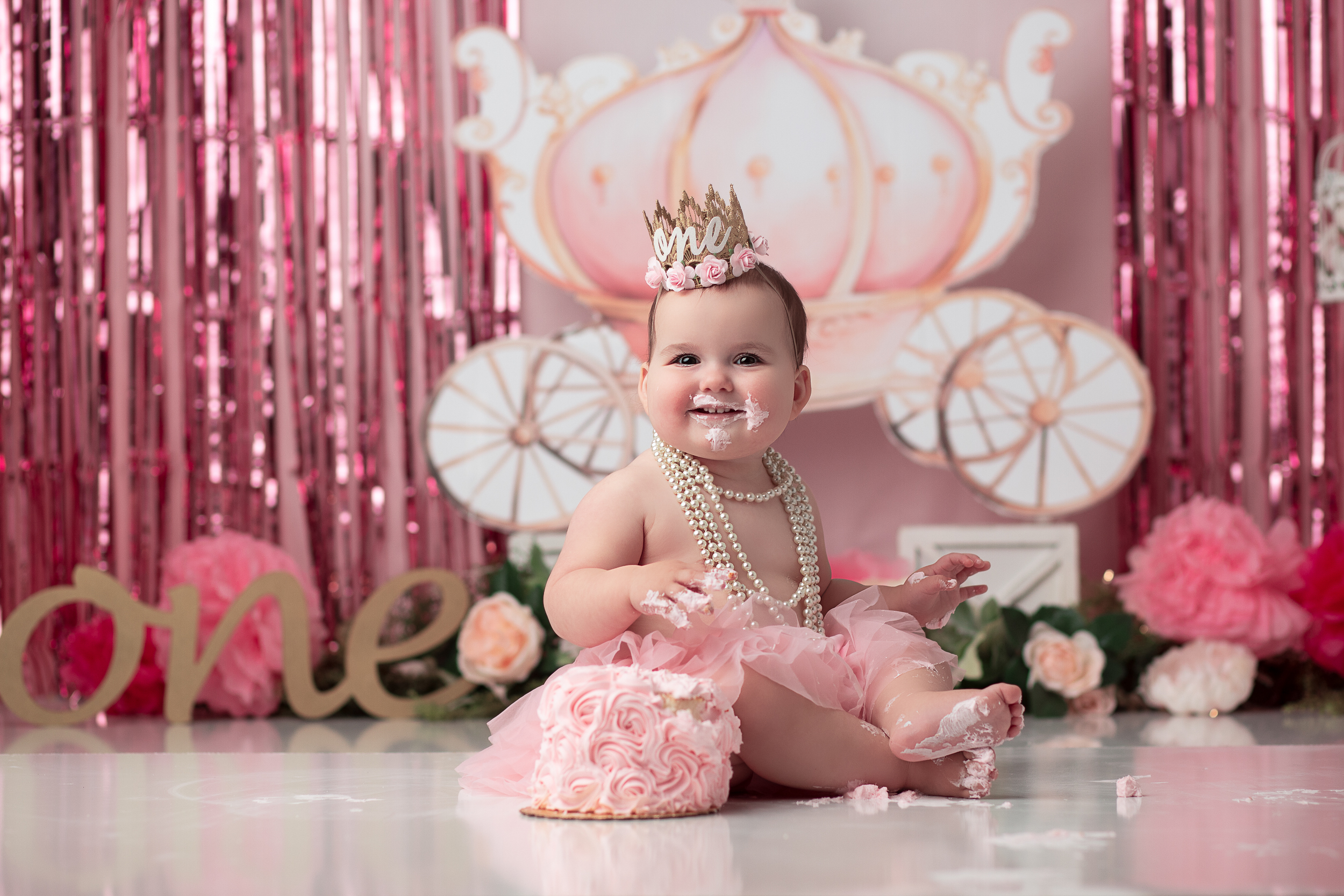 Baby Girl with a Crown and pink tutu with a cake in front of her. The backdrop is a carriage and pinks