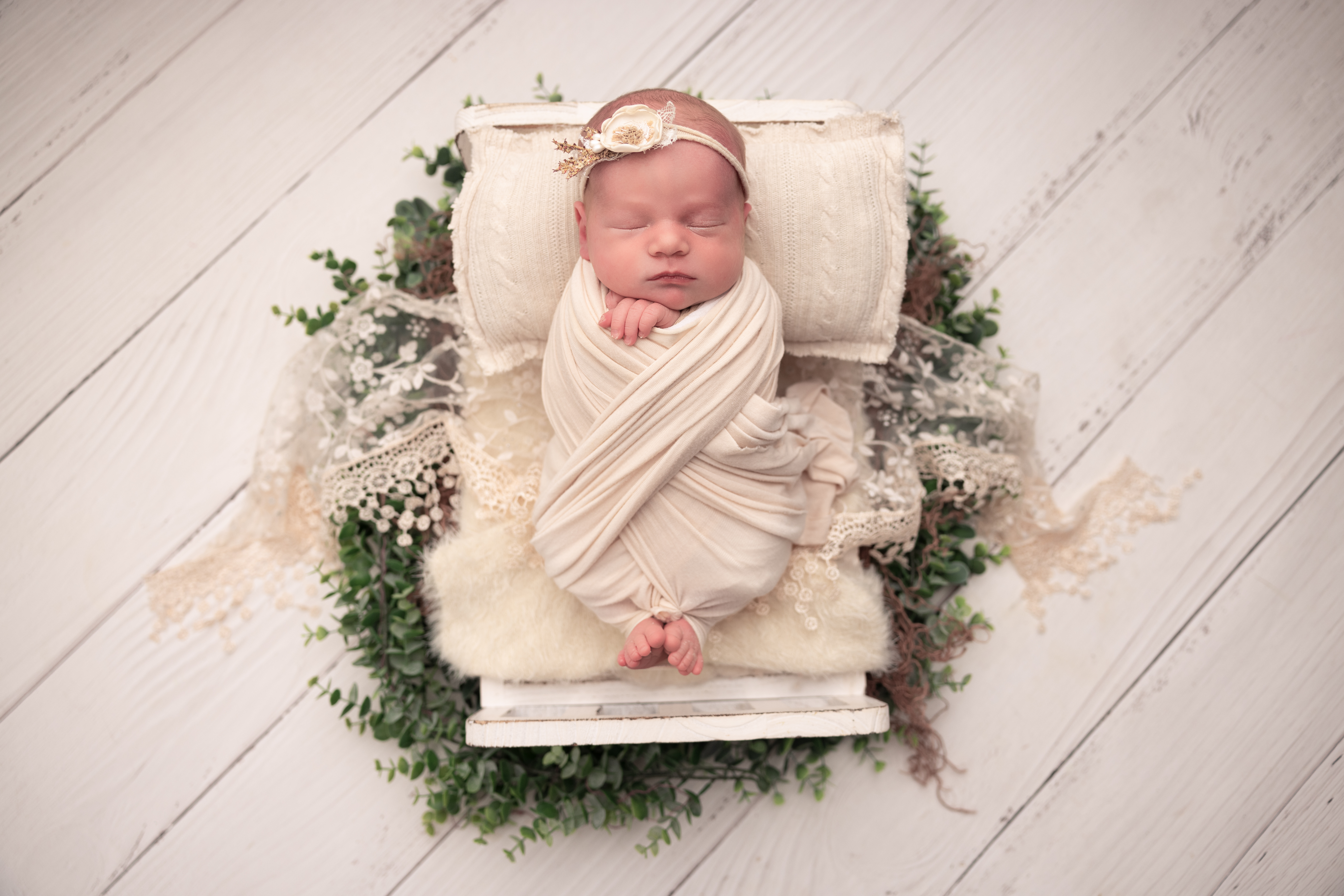 Baby girl wrapped in a cream wrap with her toes poking out lying in a baby bed with greenery