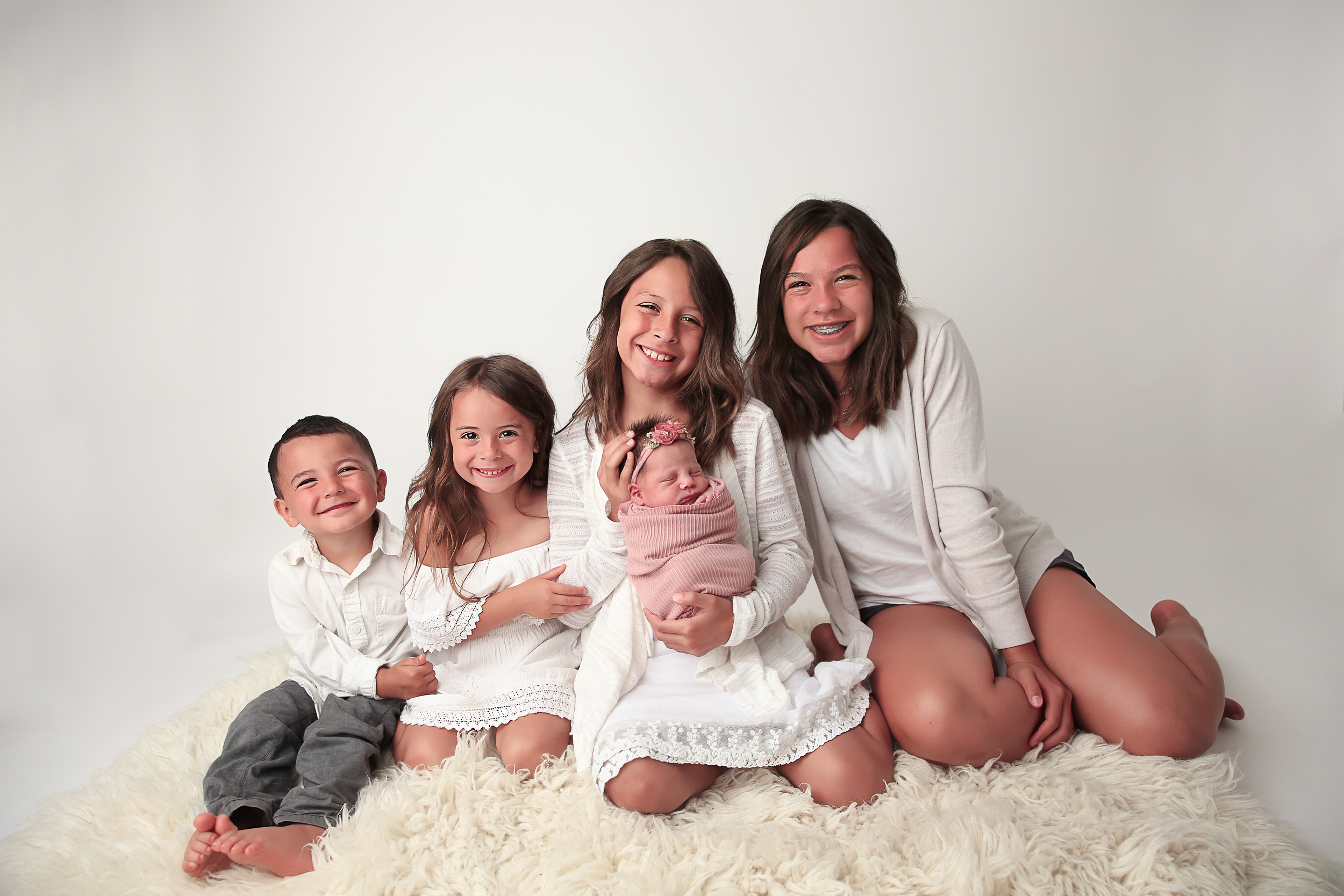 4 Siblings all sitting on a rug holding their baby sister that is wrapped in pink.