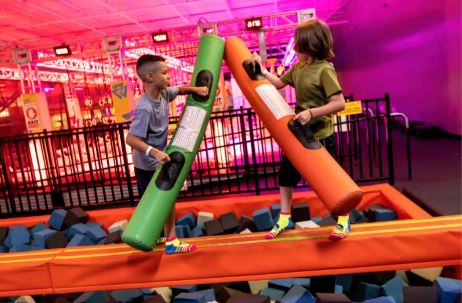 Two Boys standing on a beam with a foam pit underneath them holding a foam bar and battling. 