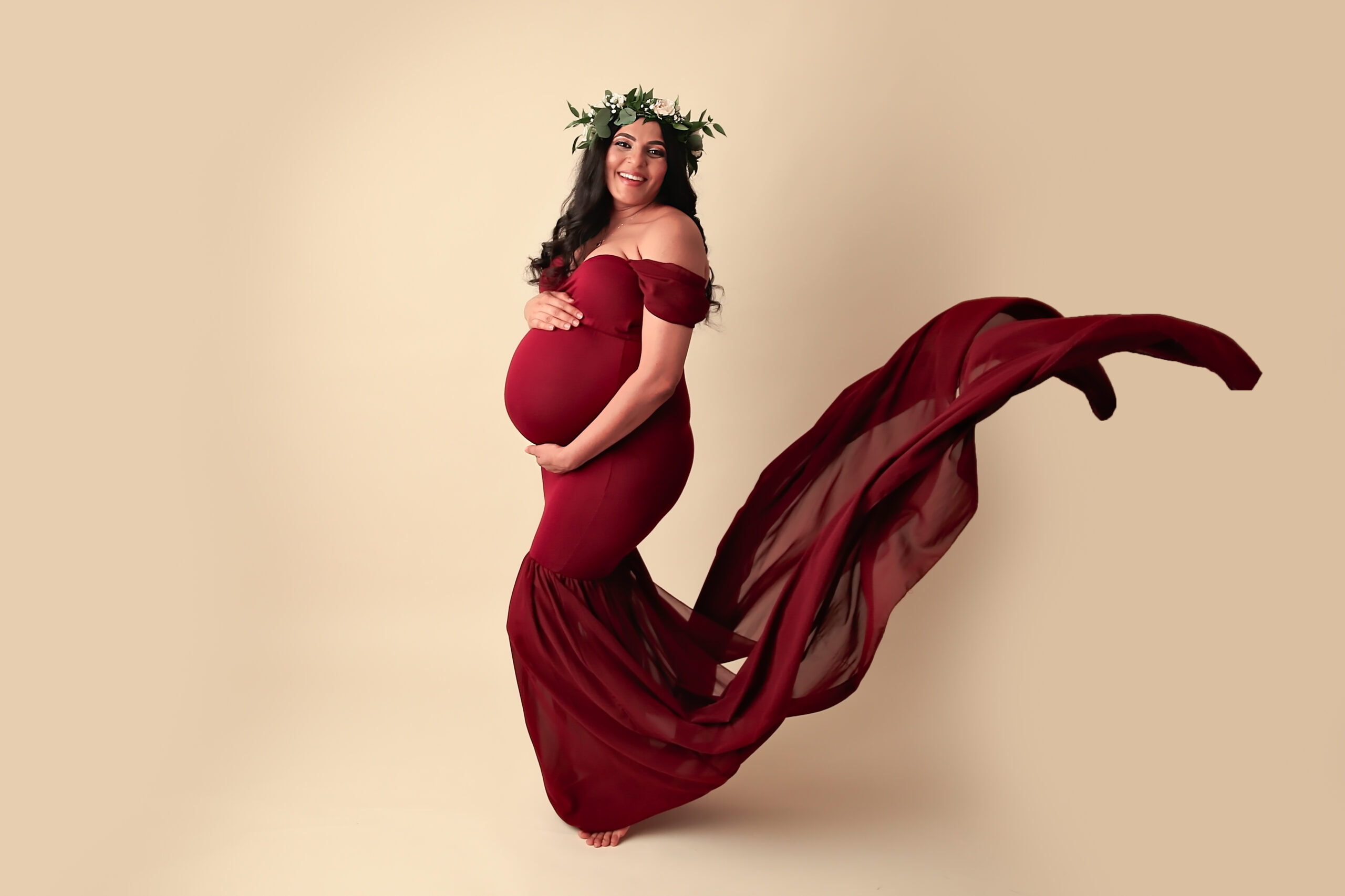 Pregnant woman with floral crown on head wearing a red maternity gown with a train flying up in the air