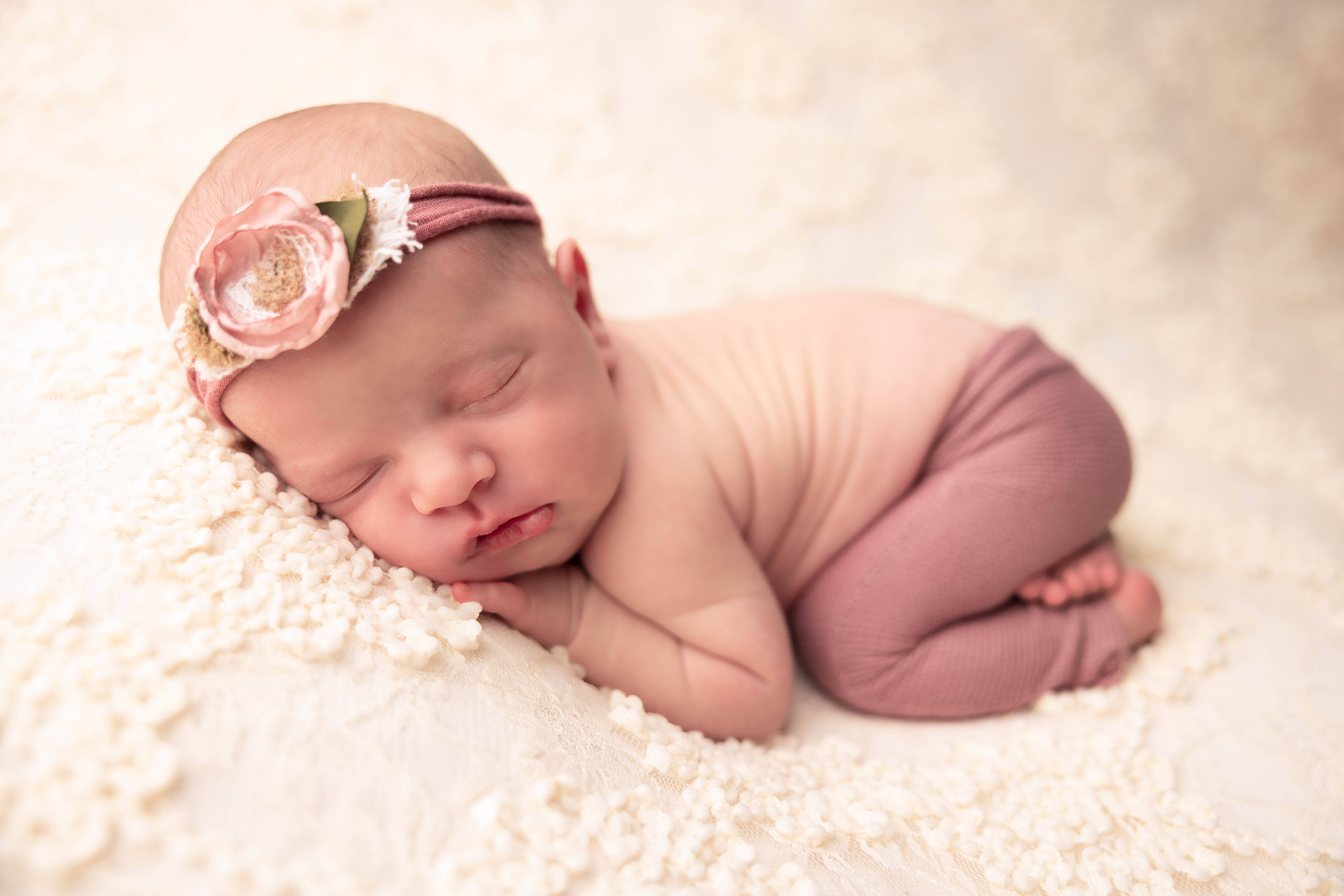Newborn baby posed with her bum up wearing pink pants and pink headband lying on a floral lace blanket