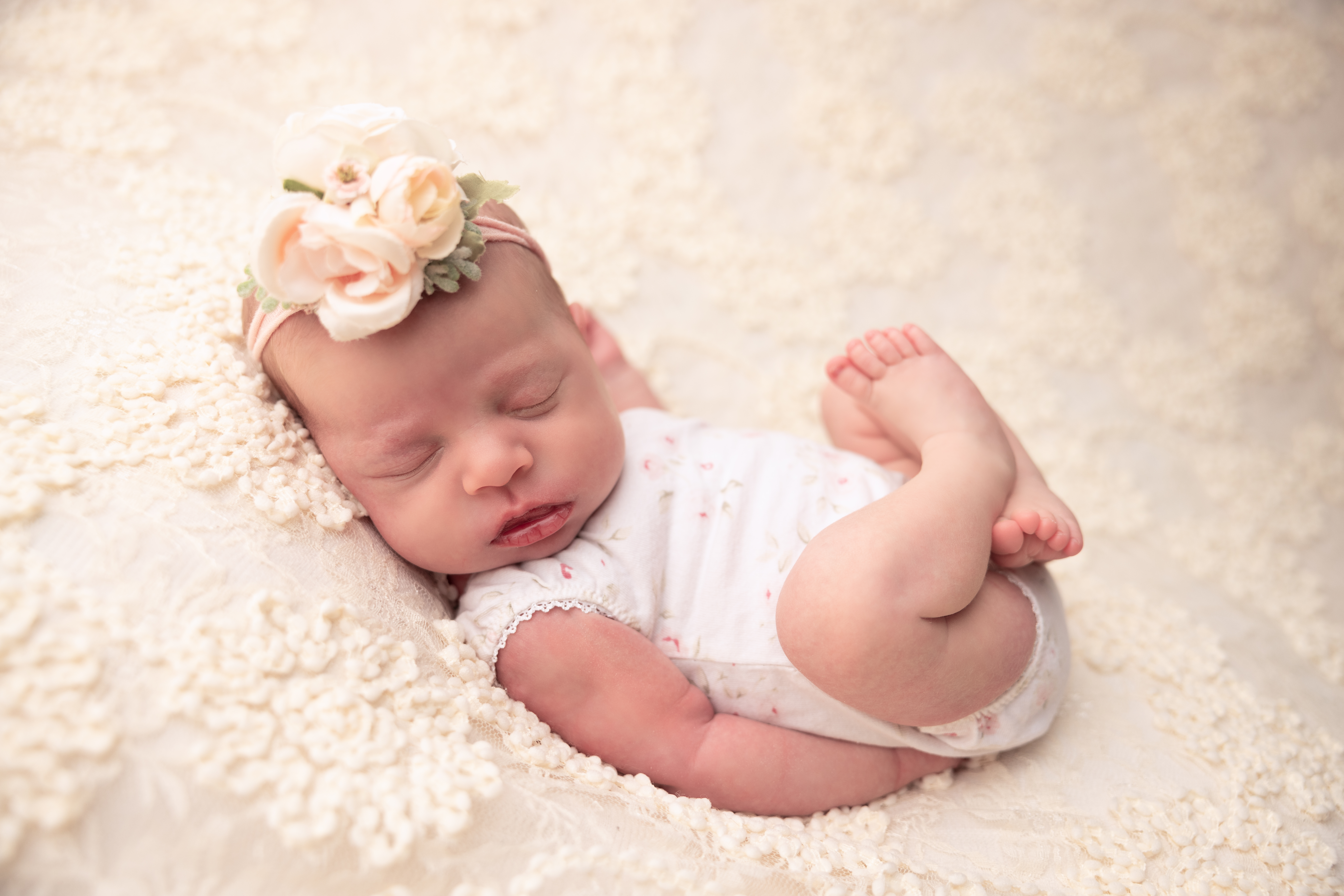 Newborn Baby in a Huck Finn Pose wearing a white romper and floral headband on a floral lace blanket