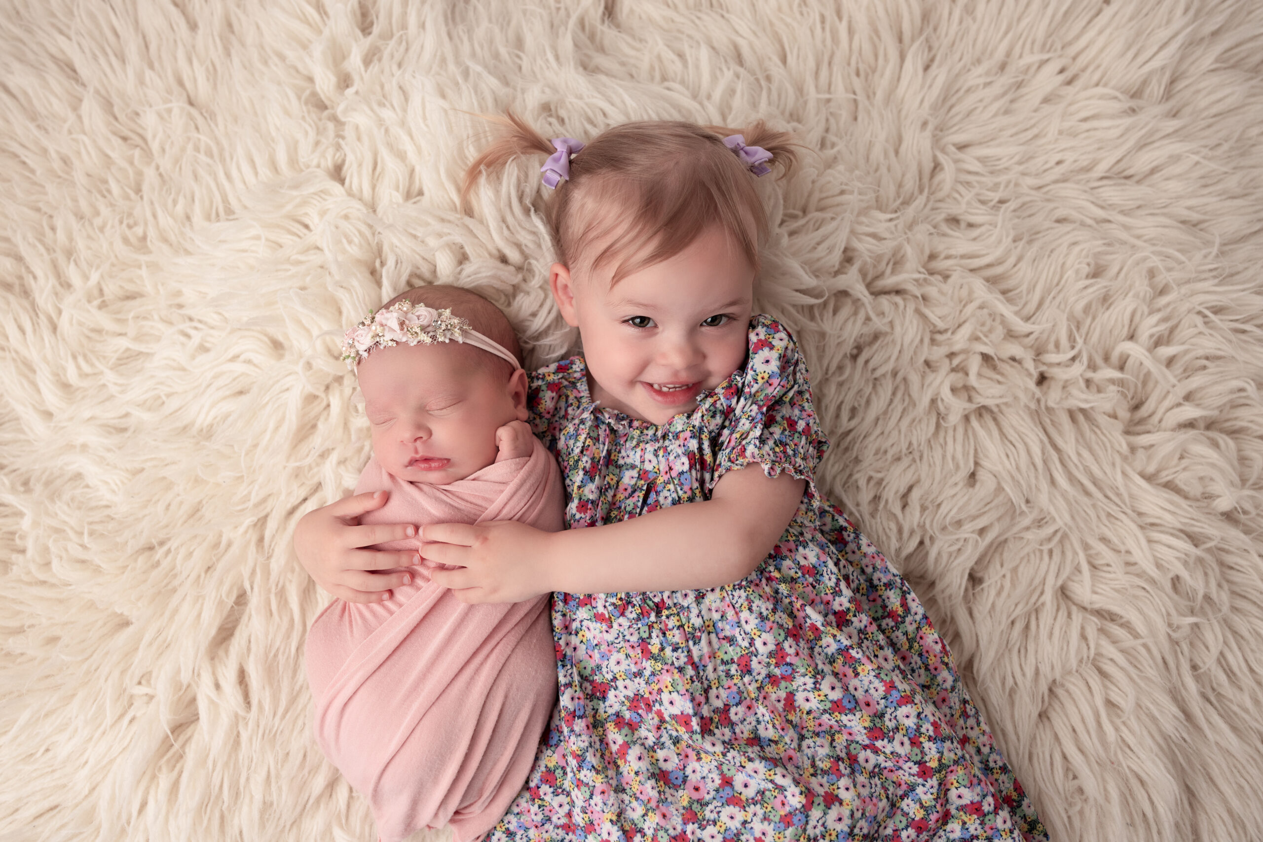 Older sibling in pig tails laying on a rug with her arms around her new baby sister wrapped in pink
