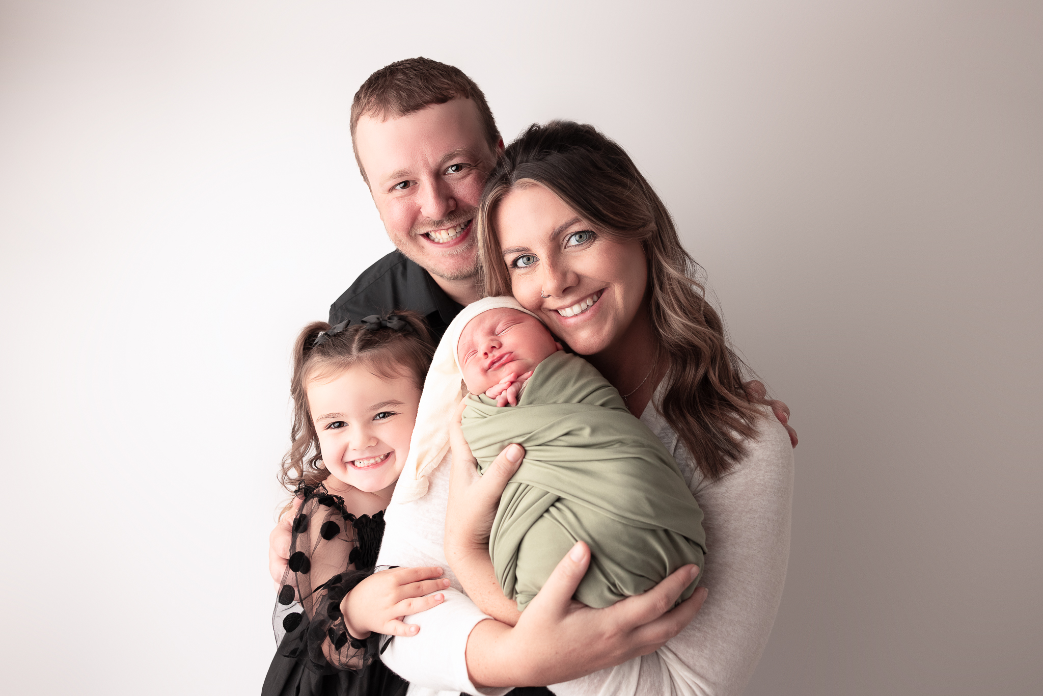 Family photo of Mom holding newborn baby, big sister giving mom a hug and dad embracing the entire family