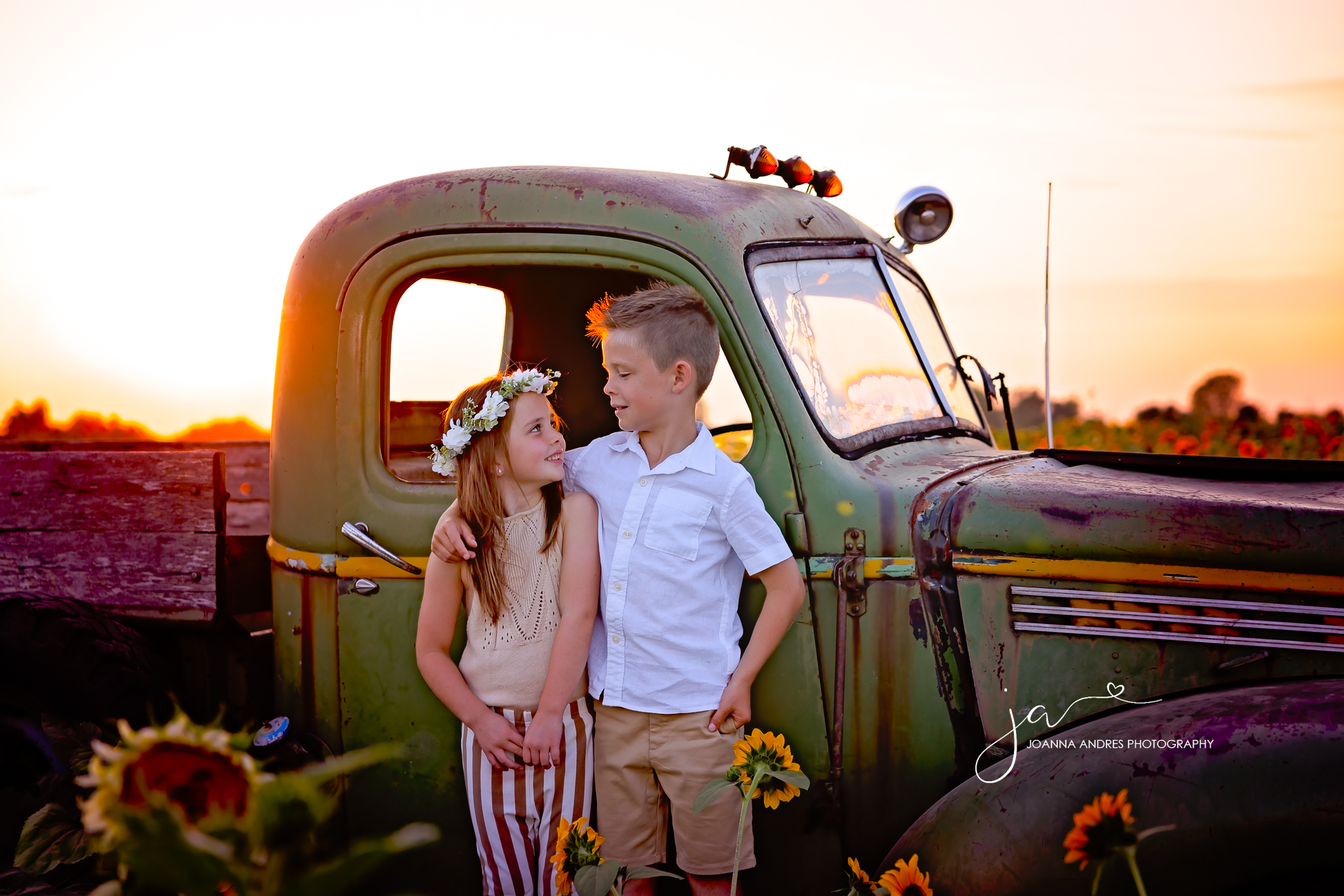 Photo of girl and boy siblings looking at each other. They are standing in front of a green old pick up truck in a middle of a sunflower field at Lynd Farm