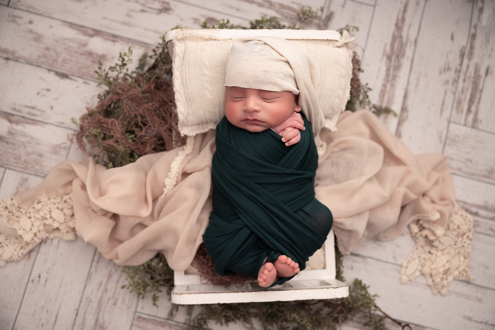 Baby in a a tiny baby bed. Swaddled in a forrest green wrap with a cream hat. 