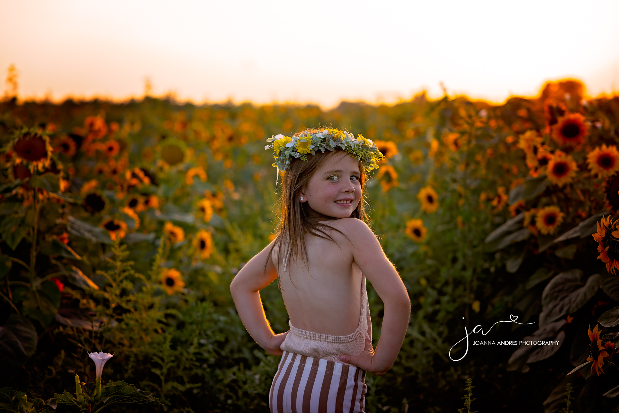 girl with her hands on her hips and you can see her back and her head is turned toward the camera smiling with a sunflower crown and with sunflowers in the backdrop