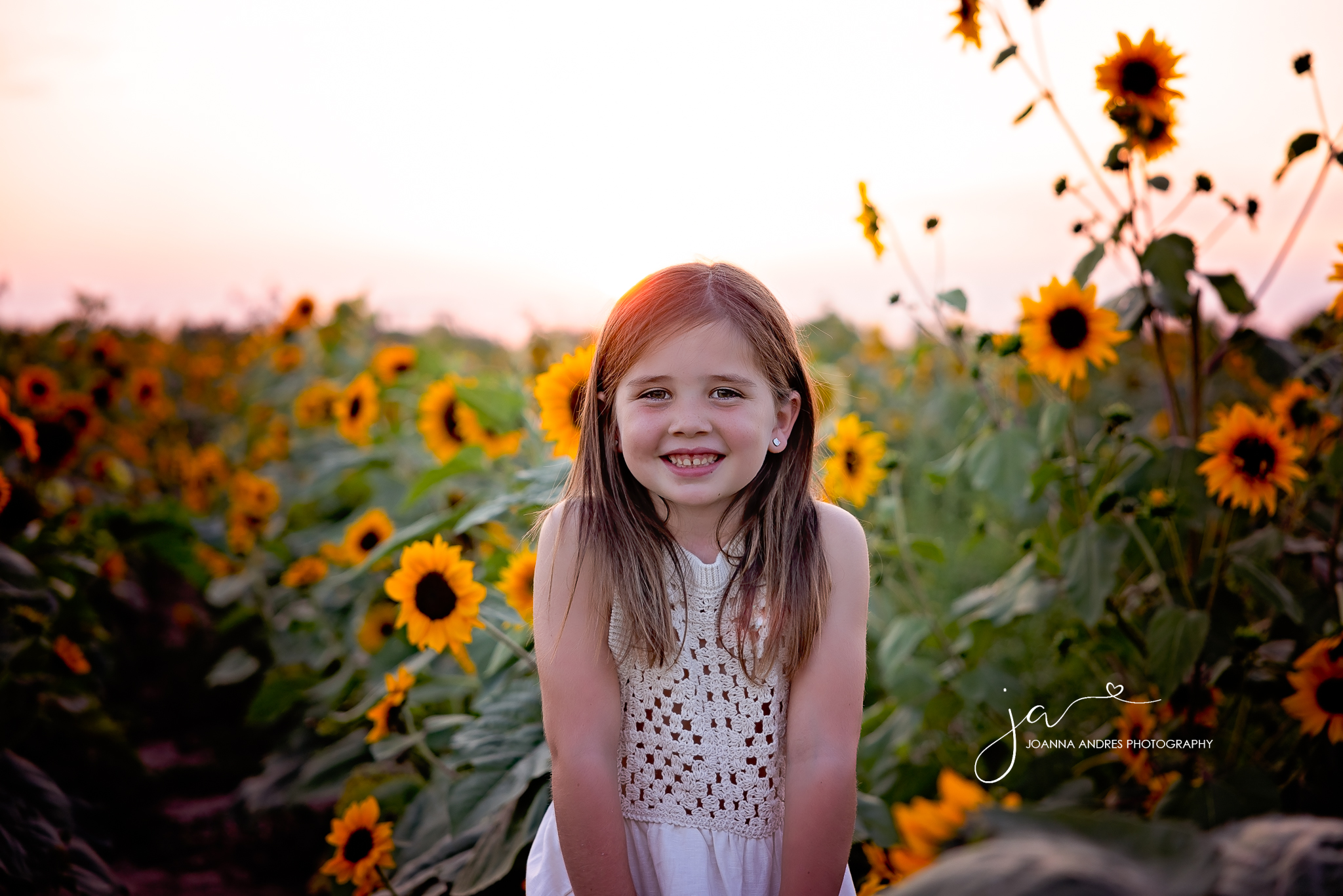 girl smiling with sunset in the backdrop and sunflowers surrounding her. 