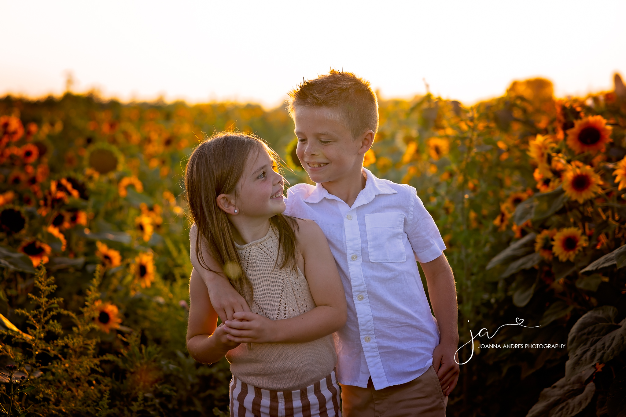 Silbings smiling and looking at eachother in the middle of a sunflower field