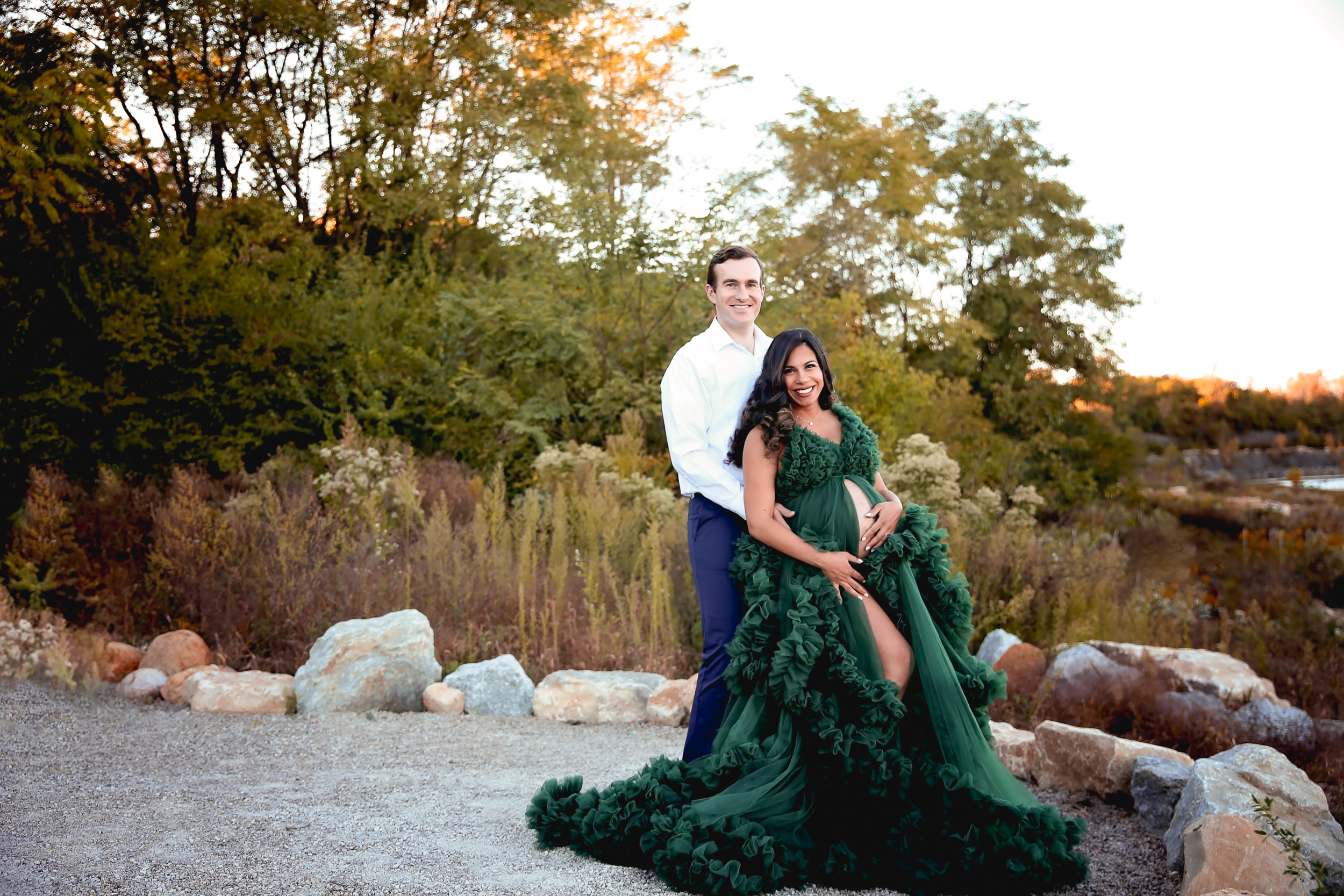 Photo of Woman in a green maternity dress showing her baby bump with her spouse behind her. Backdrop is rocks and trees. 