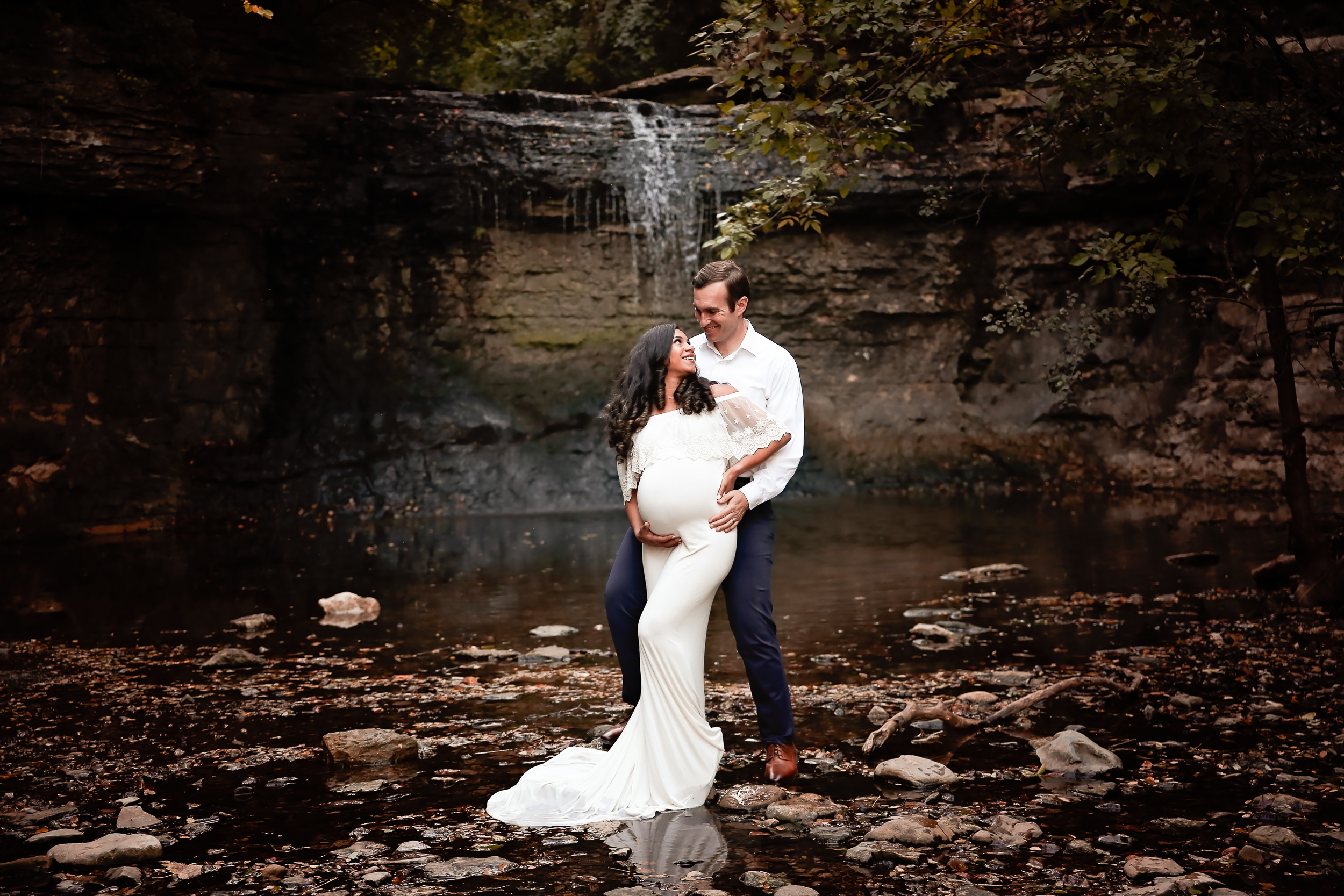 Maternity PHoto of woman in a white fitted dress looking back at her husband with a waterfall in the backdrop