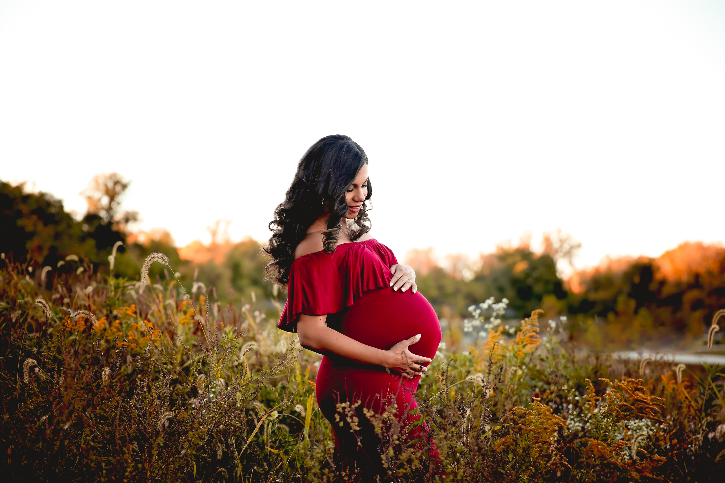 Pregnant woman in a red sleeveless dress looking down at her bump in a sea of wild flowers in the fall