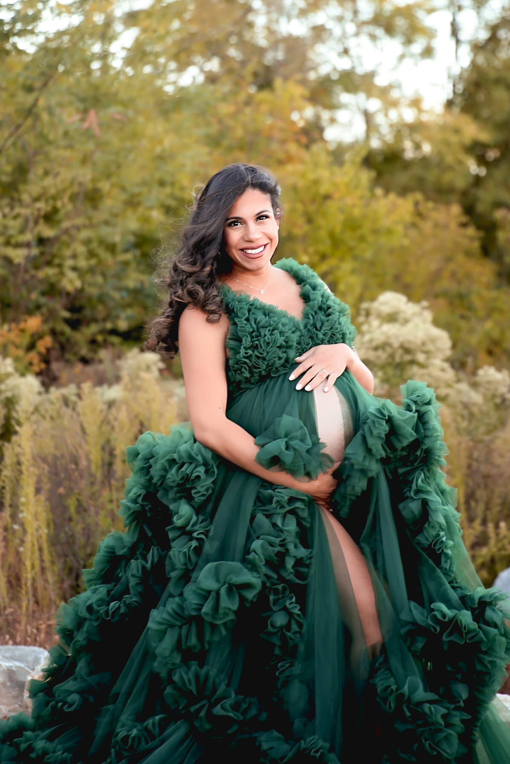 Woman is pregnant wearing a green maternity gown with her baby bump peaking out with a beautiful badkrop of quarry trails park