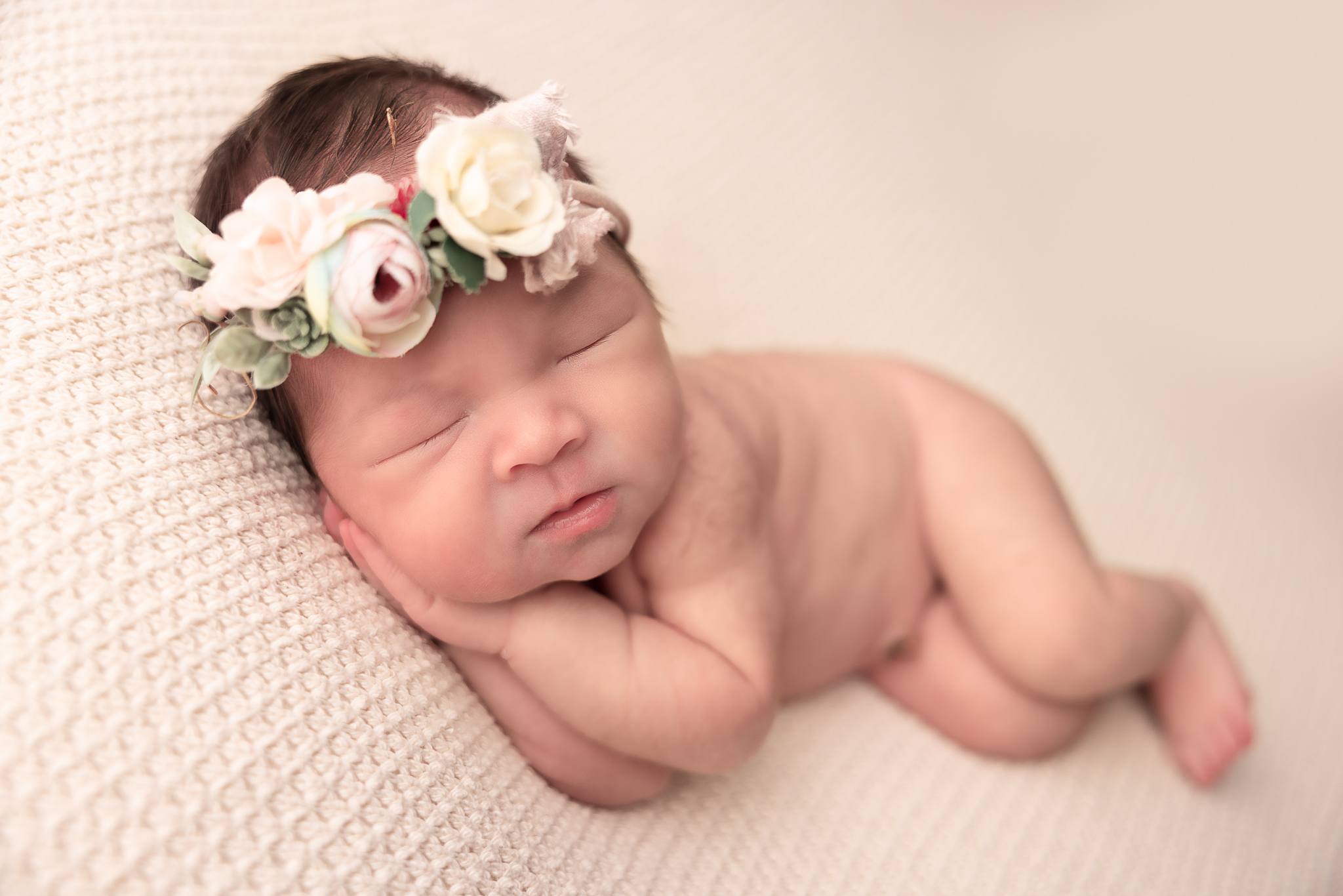 Side laying pose of baby sleeping with both hands under her cheek with floral headband.