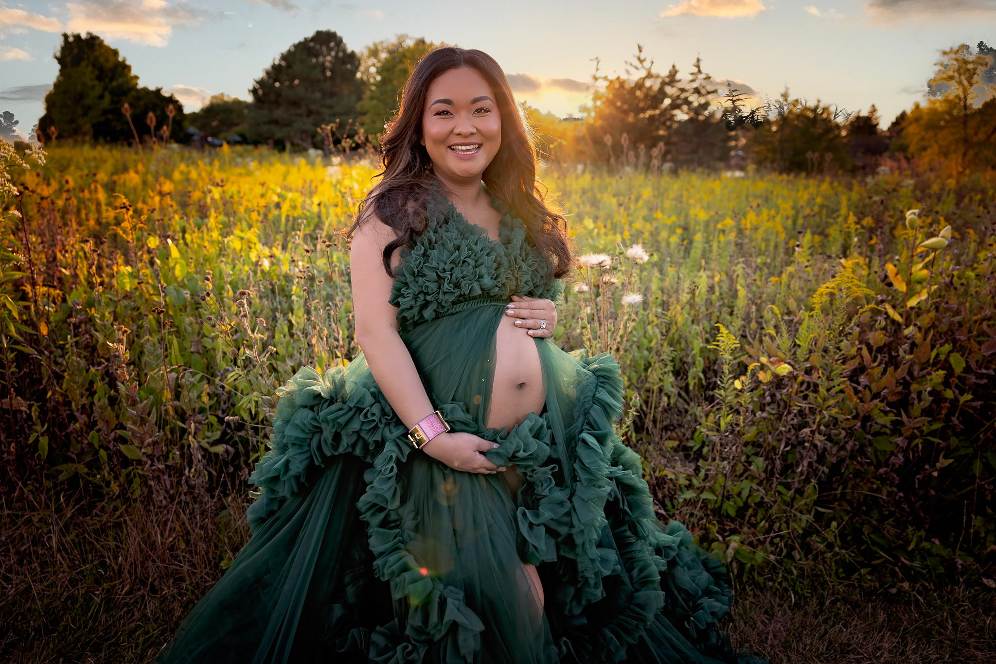 Pregnant woman wearing a green dress with her pregnant belly peaking out. The bacdkrop is long grass, wildflowers and a sunset