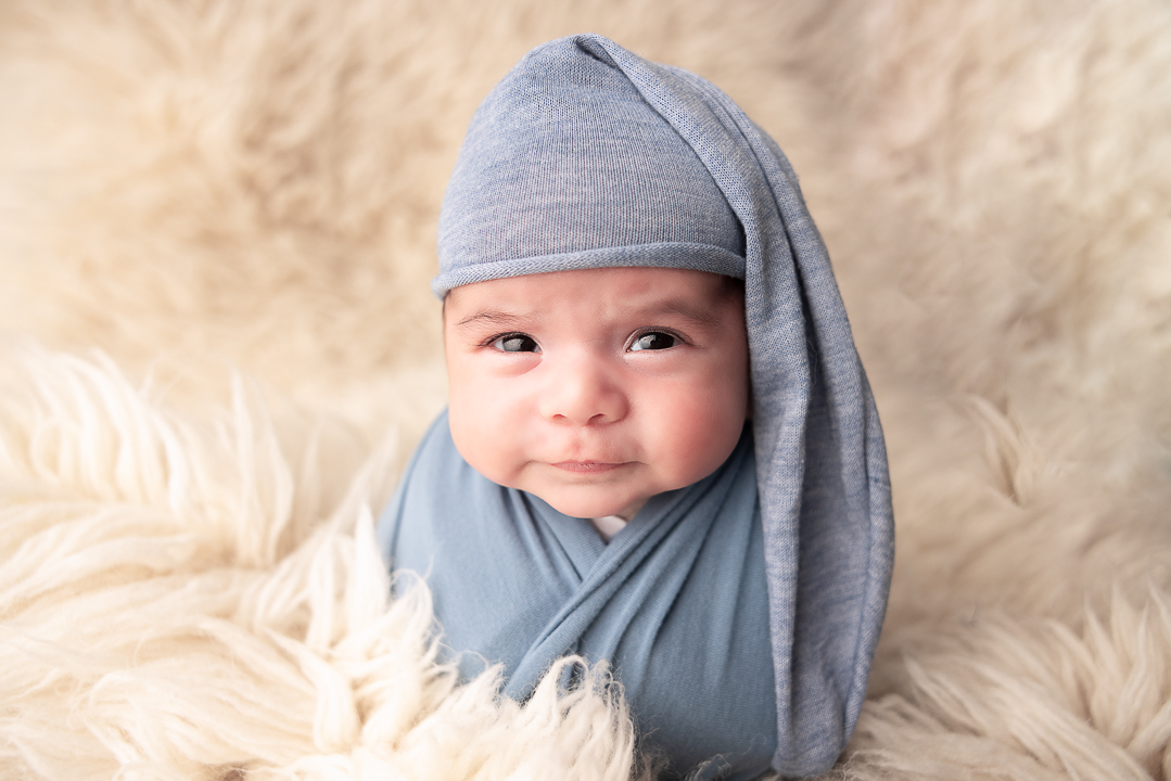 Baby boy wrapped in blue wearing a blue hat and with a cream flokati rug surrounding him