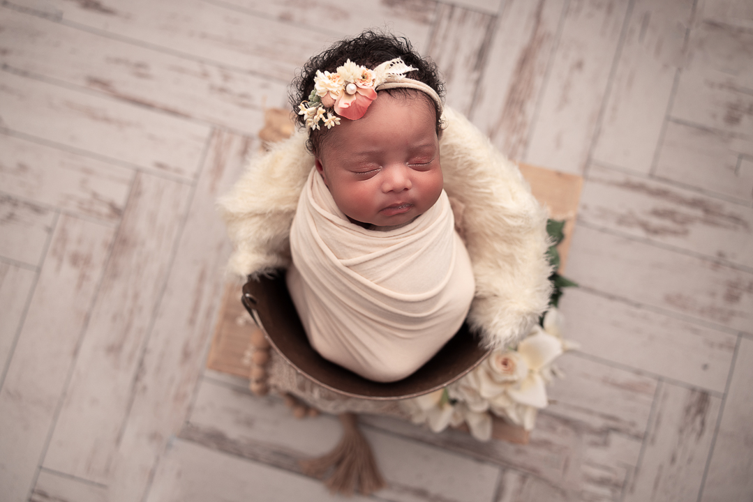 Baby Girl in a bucket wrapped in a cream wrap with a headband on her head full of curly hair