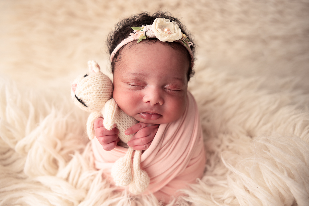 Baby girl holding a kitten lovey and she's wrapped in a pink wrap wearhing a floral headband on her black curly hair