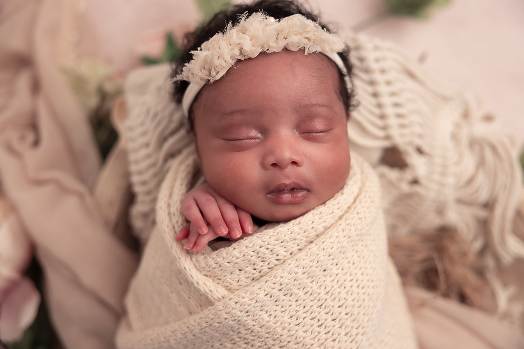 Close up of baby's face that is wrapped in a cream textured blanket wearing a cream floral headband