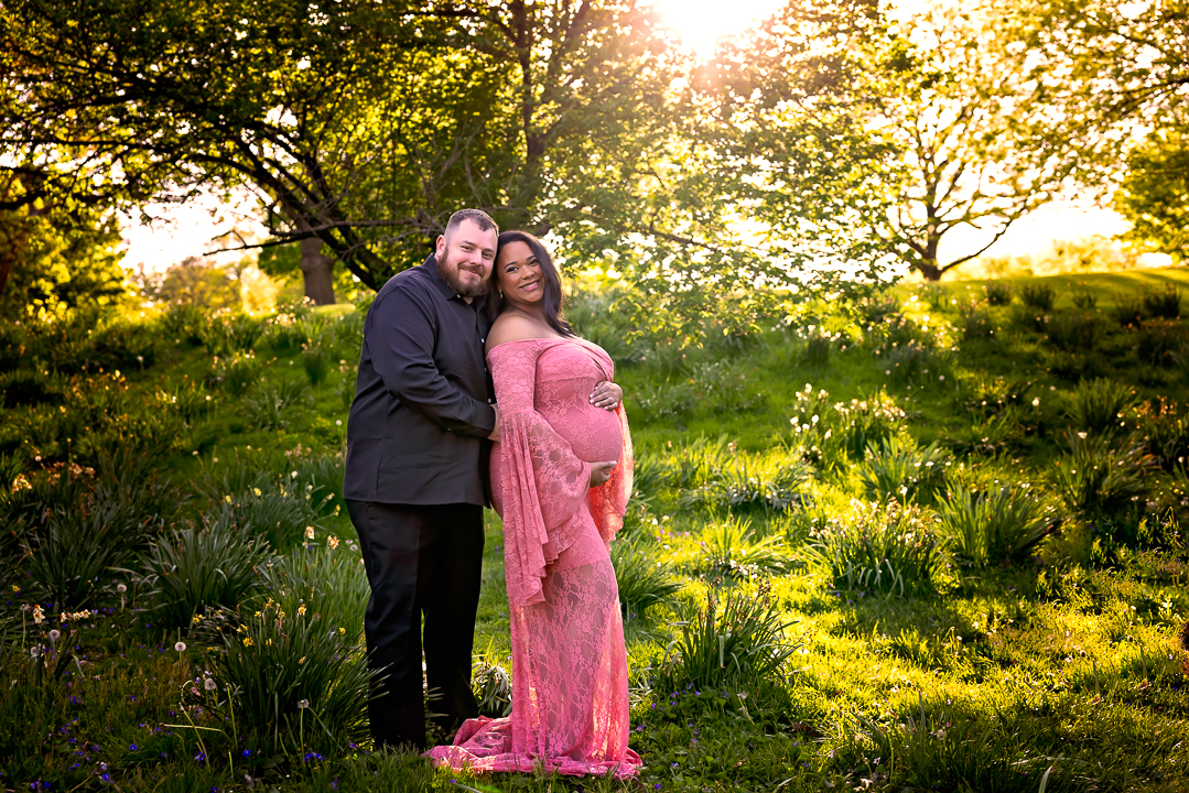 Expecting Mom wearing a pink maternity gown with her husband in a beautiful greenery setting and sunset