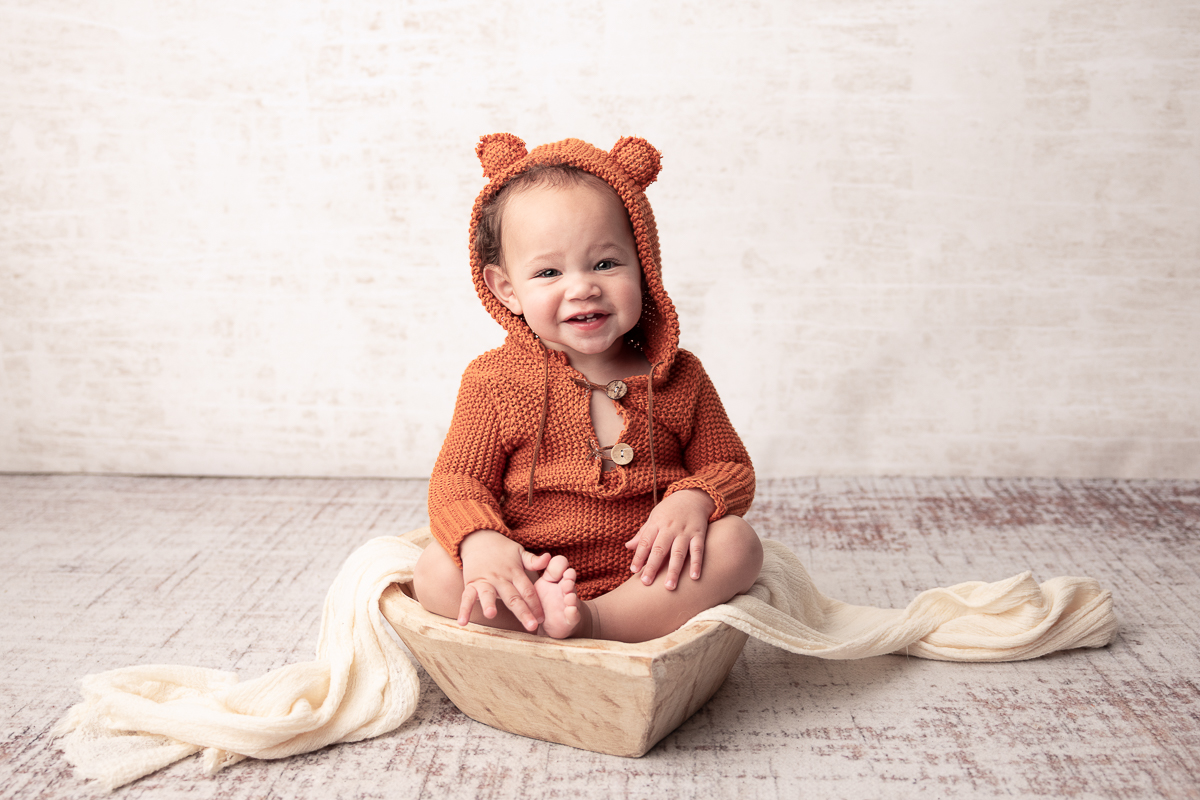 Clean portriat of baby boy in an orange teddy knitted romper sitting in a heart shaped prop for one-year photos, taken before the cake smash
