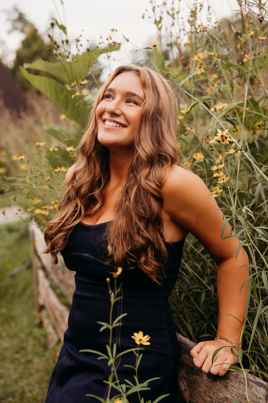 senior high school girl smiling looking up while leaning against a fence with yellow flowers and greenery behind her
