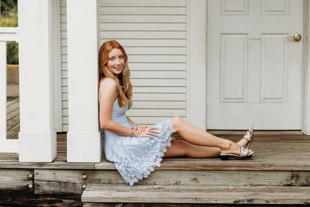Senior High School Girl sitting on a porch with white door and siding looks like a farmhouse behind her